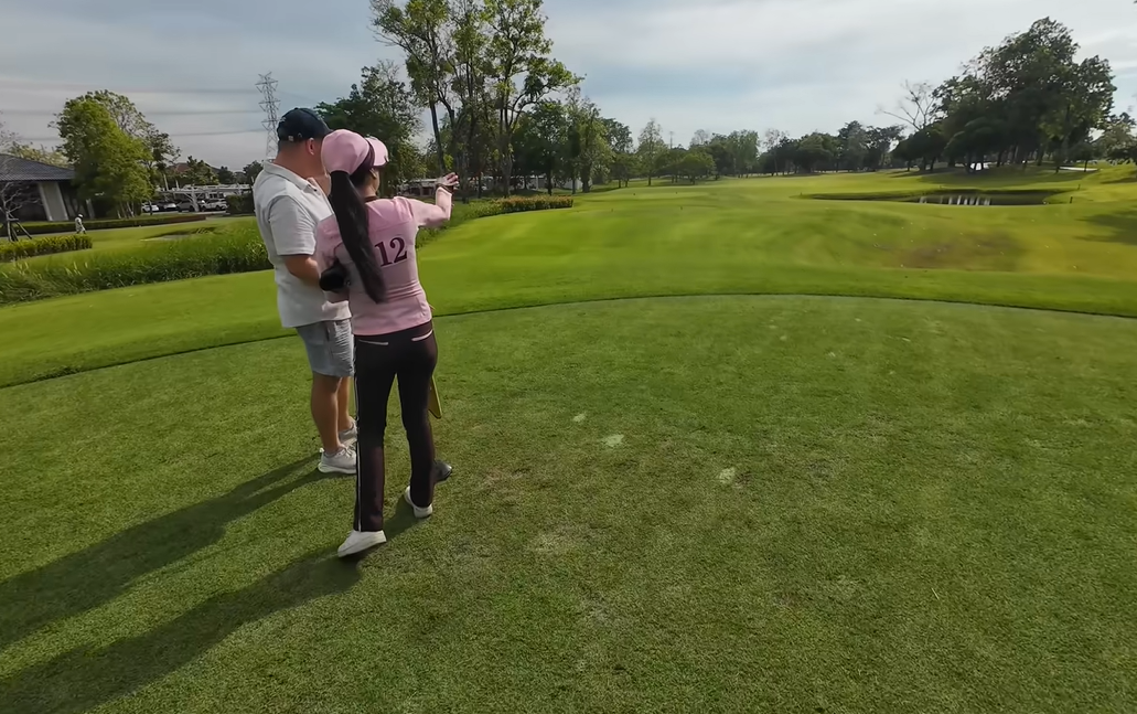 Thai golf caddie in a pink uniform pointing down the fairway to assist a male golfer on a sunny daytime golf course.