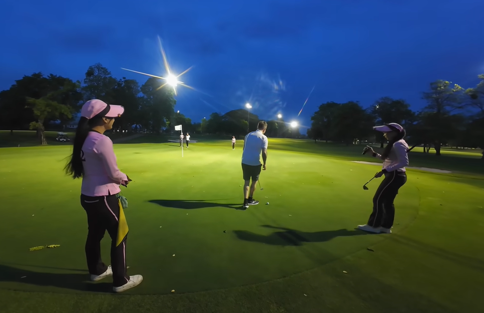 Male golfer putting on a floodlit green at night, accompanied by two Thai caddies in pink uniforms holding the flag and a club.