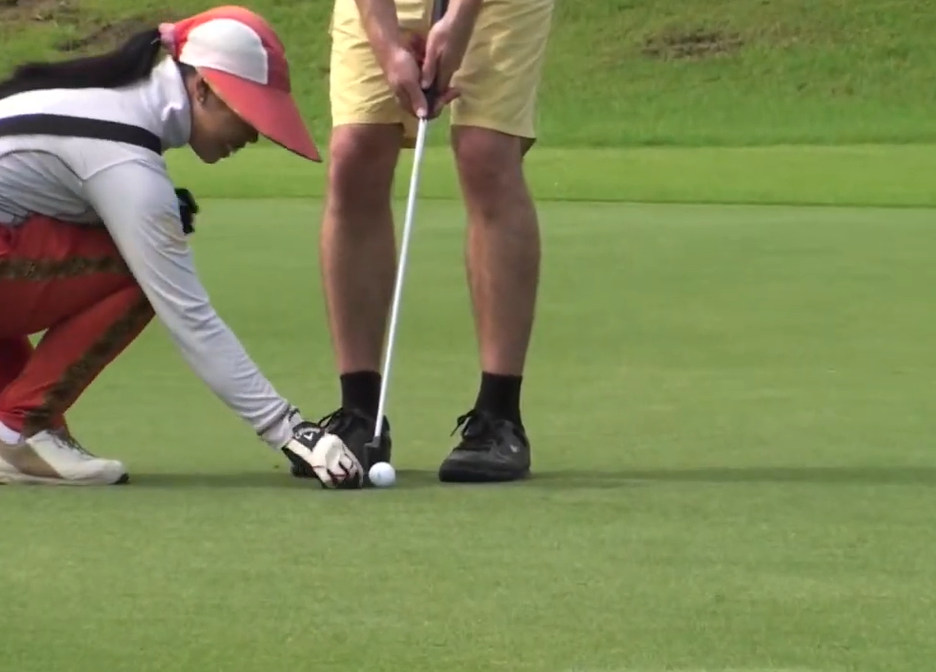 Close-up of a Thai golf caddie in a red and white visor crouching to place a golf ball on the putting green next to a golfer's putter.